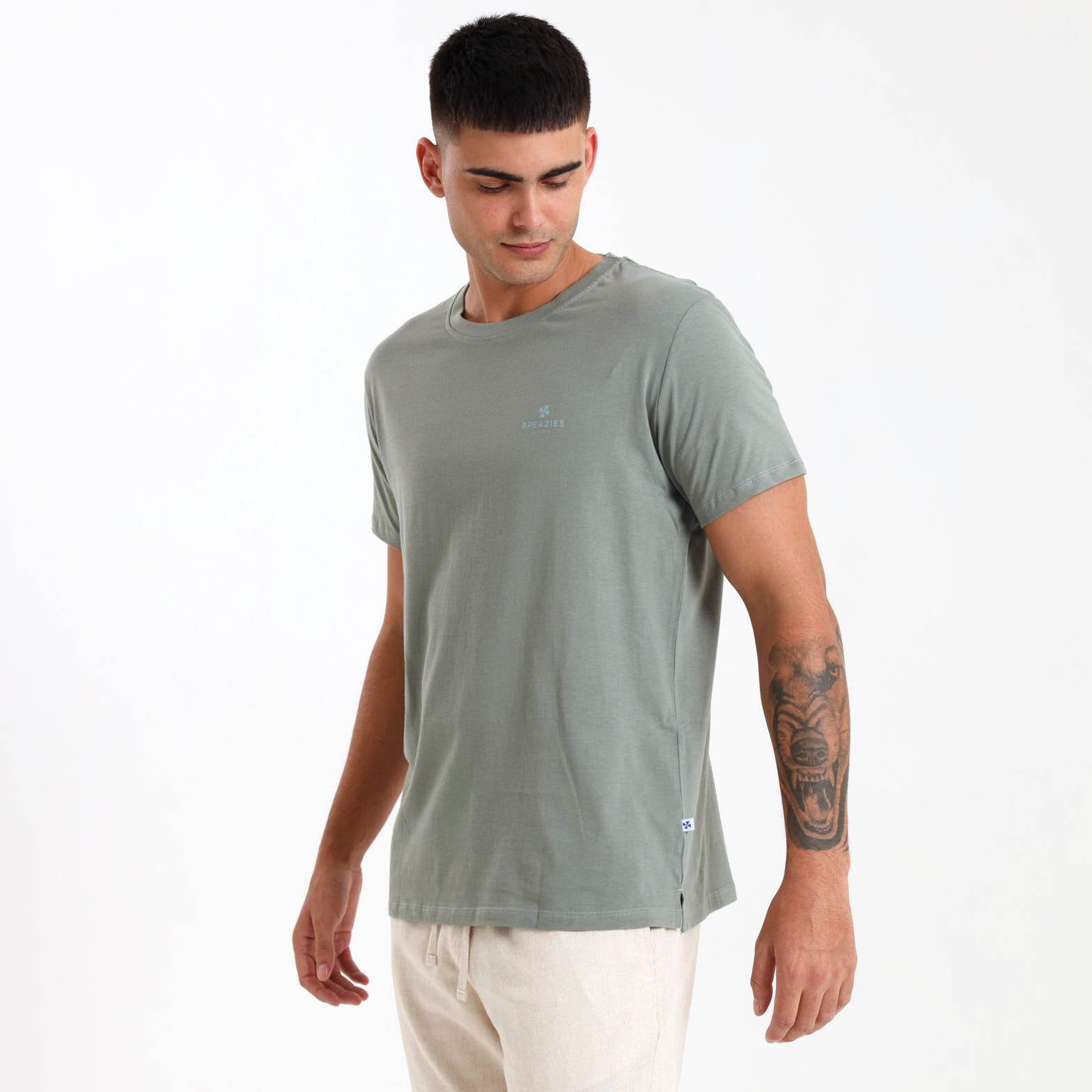 A male model wearing a sage green cotton t-shirt with subtle printed branding on the front left chest area, designed in South Africa.