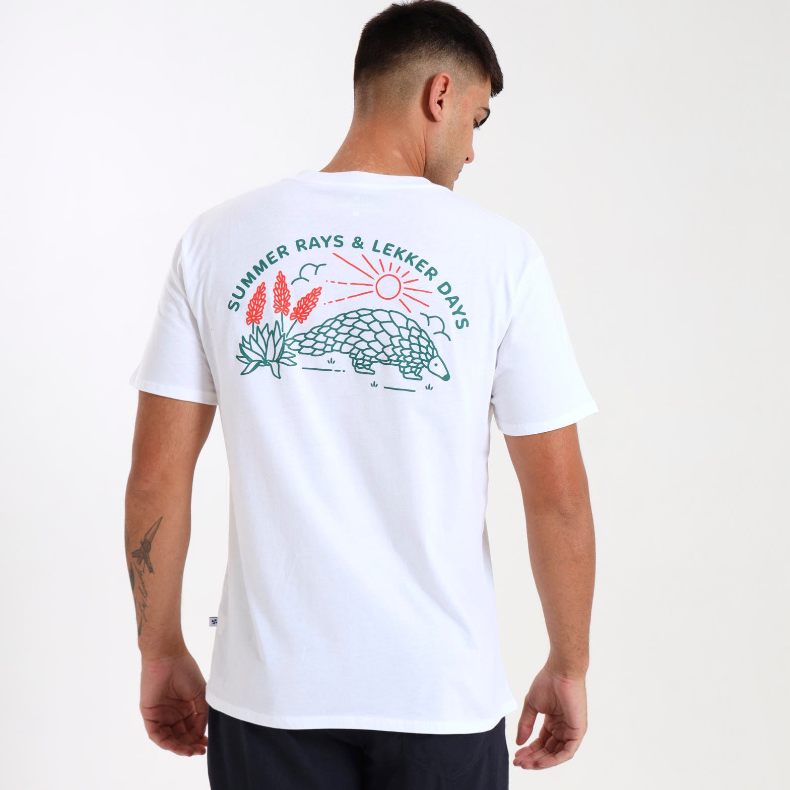A person wearing a white crew neck t-shirt with a graphic print on the back, featuring a pangolin and text saying "summer rays & lekker days".