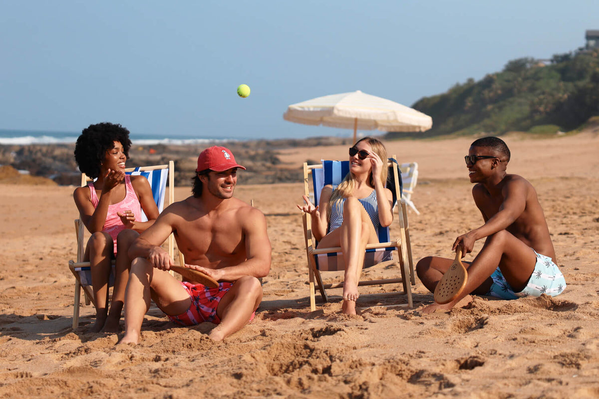 Four people sitting in beach chairs wearing Breazies swimwear on a sandy beach under a blue sky, with a white umbrella, ocean, and green hillside in the background.