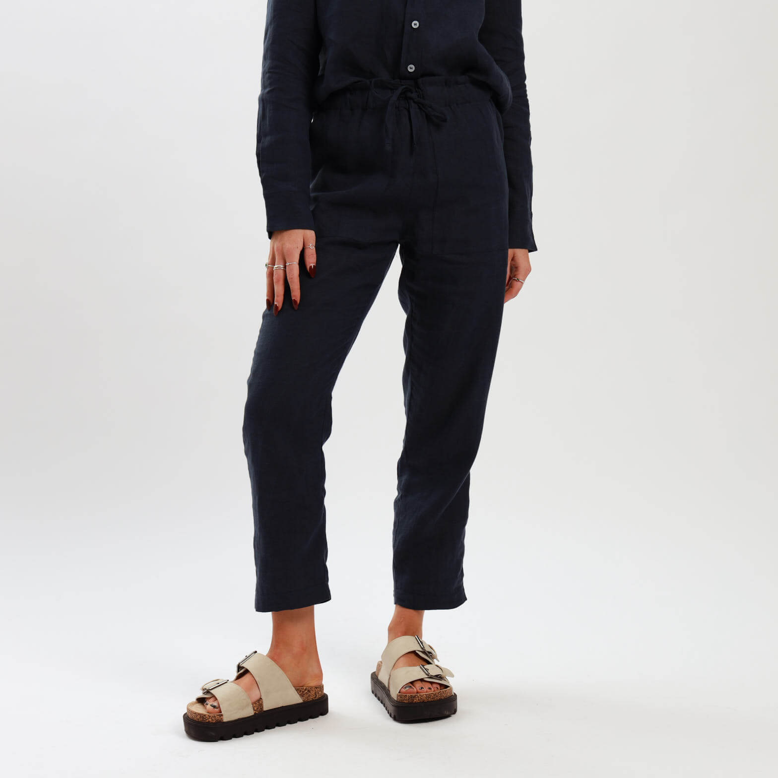 A model wearing navy high-waisted linen pants with an elasticated waistband and drawstrings, paired a matching linen shirts and tan sandals.