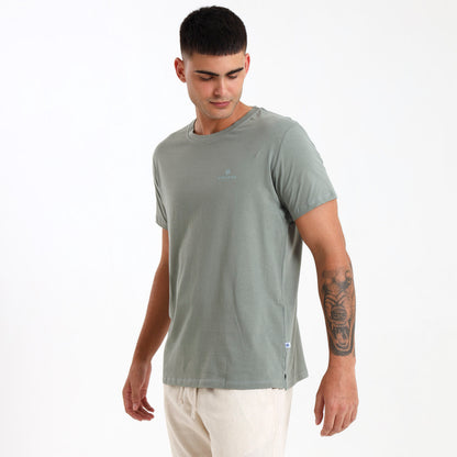 A male model wearing a sage green cotton t-shirt with subtle printed branding on the front left chest area, designed in South Africa.