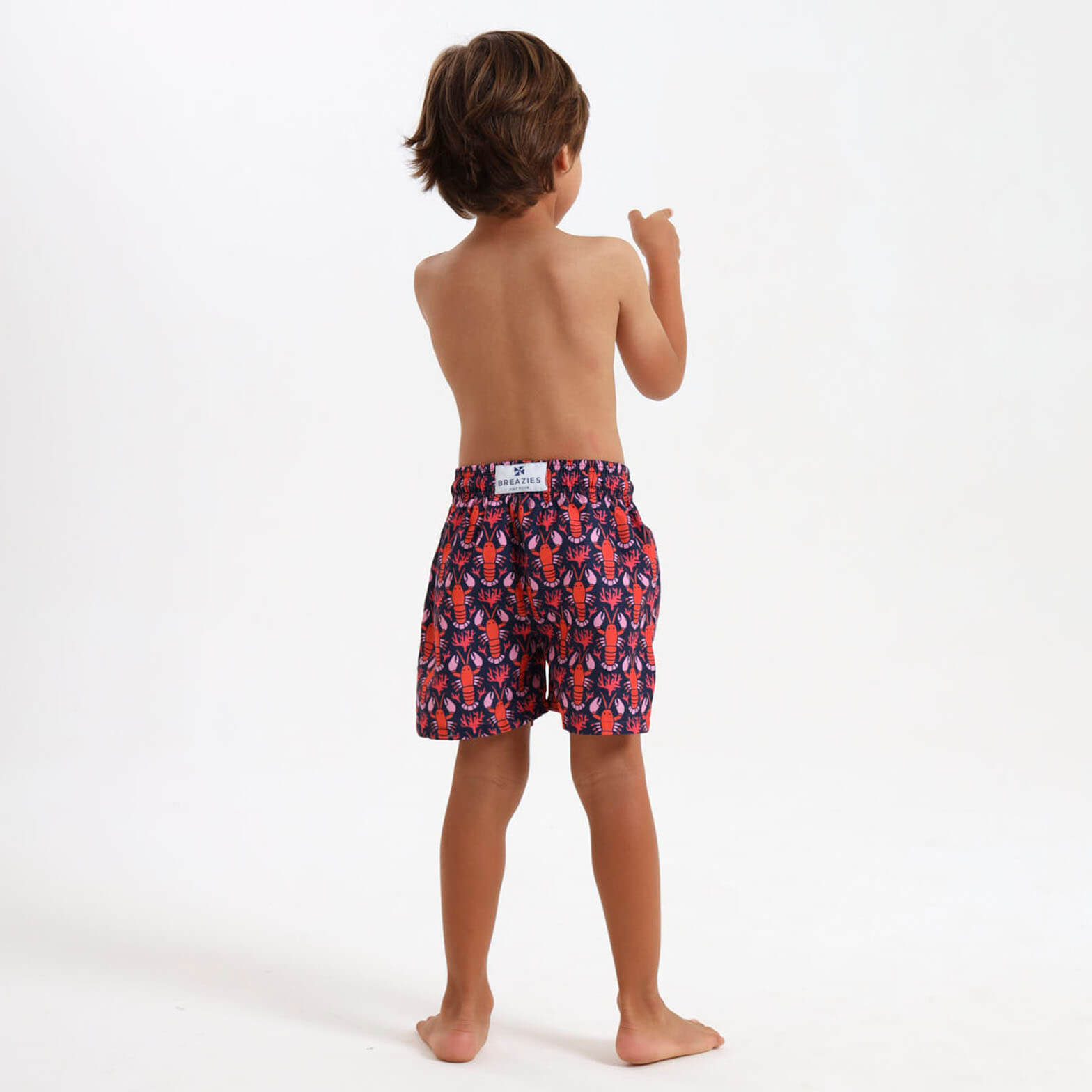 Young boy wearing a pair of navy swim shorts with orange lobster design on them.