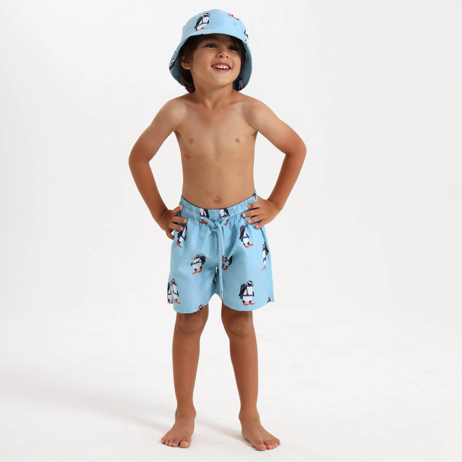 Boy wearing blue swim shorts with penguin design and a matching bucket hat.