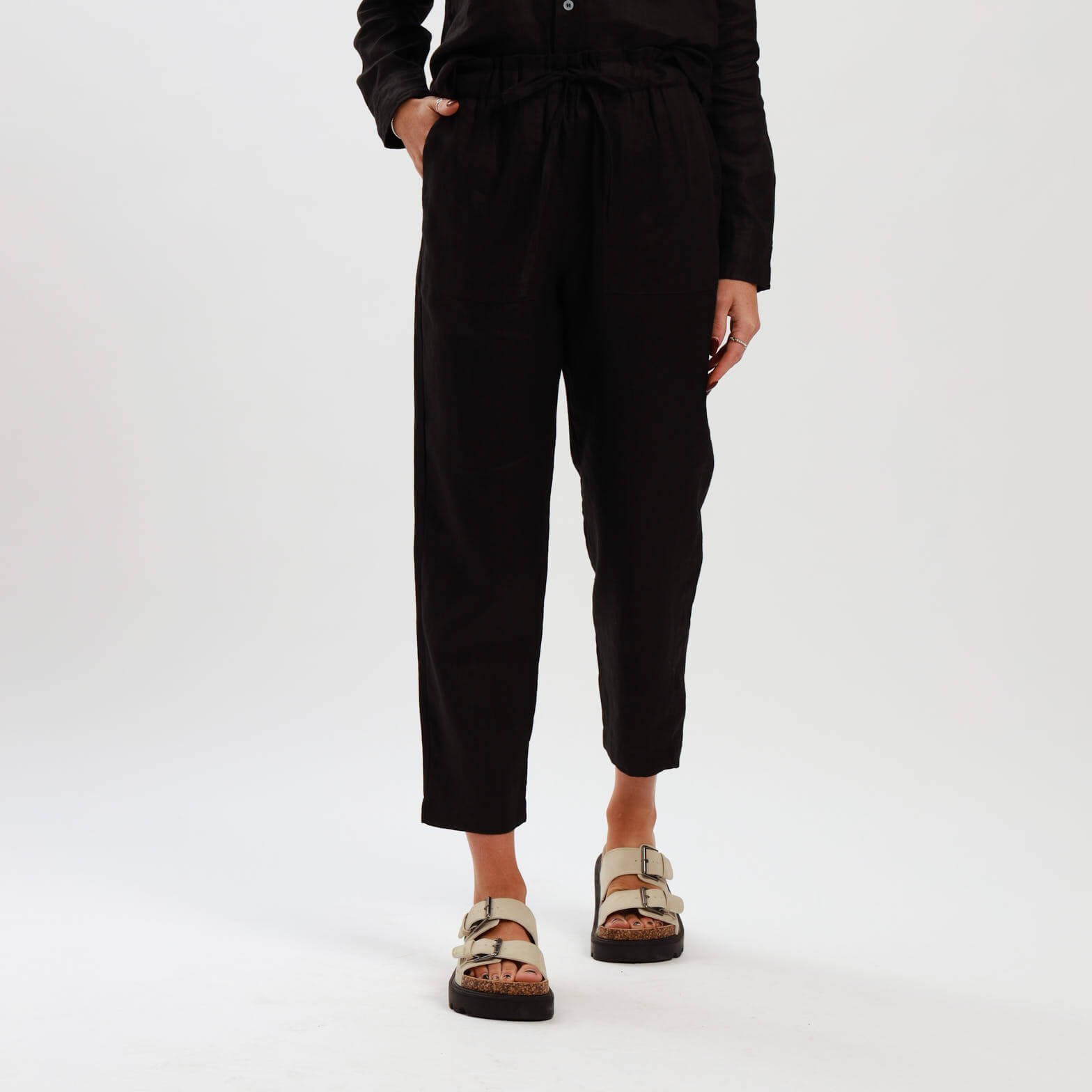 A model wearing black high waisted ladies linen pants with an elasticated waistband and tapered ankle length leg, standing against a neutral background.