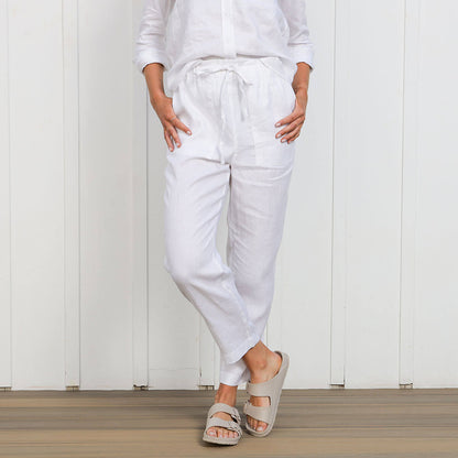 A woman standing in front of a white background wearing high waisted white linen pants with an elasticated waistband and tapered ankle length leg, along with a matching white linen long sleeve top.