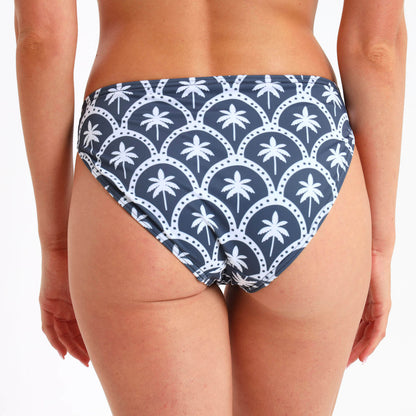 Steel blue bikini bottom with a slightly cheeky bum and an all over white palm print  being worn by a woman.